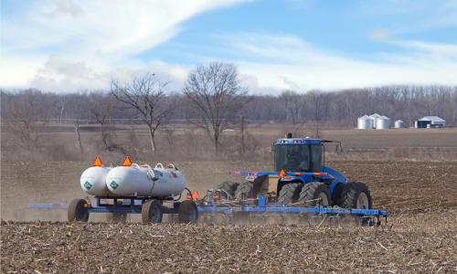 photo of a tractor in a turned-over field pulling a trailer with two tanks of anhydrous ammonia fertilizer that is being sprayed on the field; farm buildings and silos are in the distance